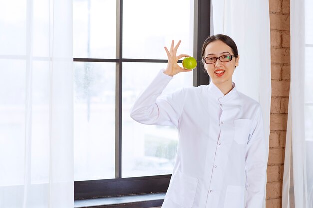 Junge Frau mit Brille im Laborkittel, die grünen Apfel in der Nähe des Fensters hält.