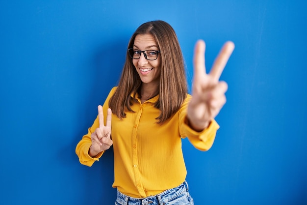 Junge Frau mit Brille steht vor blauem Hintergrund und blickt lächelnd in die Kamera, während sie mit den Fingern das Siegeszeichen Nummer zwei macht