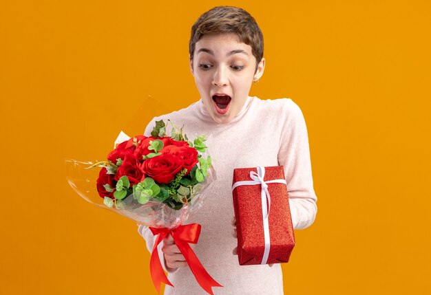 junge Frau mit dem kurzen Haar, das Strauß der roten Rosen und ein Geschenk hält, das erstauntes und überraschtes Valentinstagkonzept steht, das über orange Wand steht