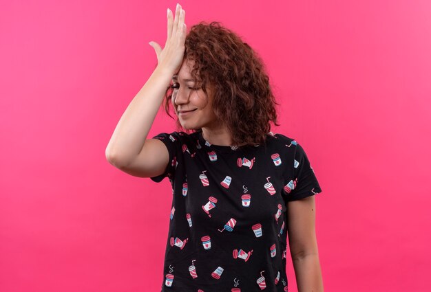 Junge Frau mit dem kurzen lockigen Haar, das mit Hand auf Kopf für Fehler steht, vergessen, schlechtes Gedächtniskonzept, das über rosa Wand steht