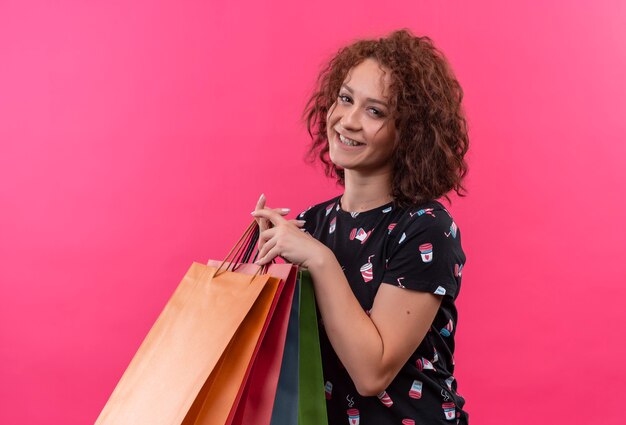 Junge Frau mit dem kurzen lockigen Haar, das Papiertüten hält, die fröhlich glücklich und positiv stehen über rosa Wand lächeln