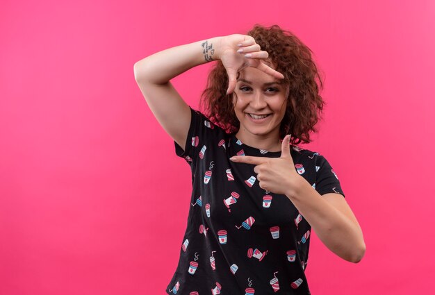 Junge Frau mit dem kurzen lockigen Haar, das Rahmen mit den Fingern macht, die durch diesen Rahmen schauen, der fröhlich über rosa Wand stehend lächelt