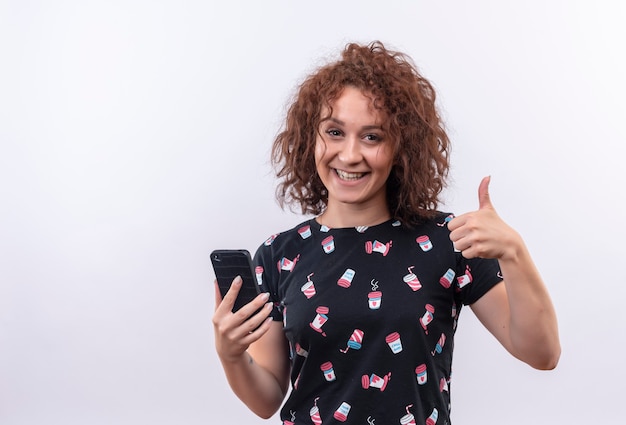 Junge Frau mit dem kurzen lockigen Haar, das Smartphone hält, das lächelnd fröhlich zeigt Daumen hoch, die über weißer Wand stehen