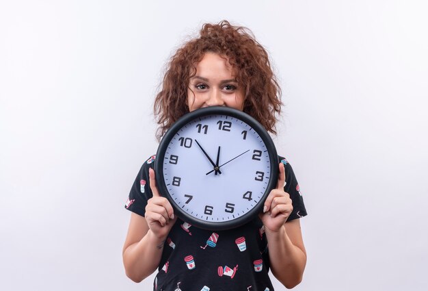 Junge Frau mit dem kurzen lockigen Haar, das Wanduhr glücklich lächelnd steht über weißer Wand hält