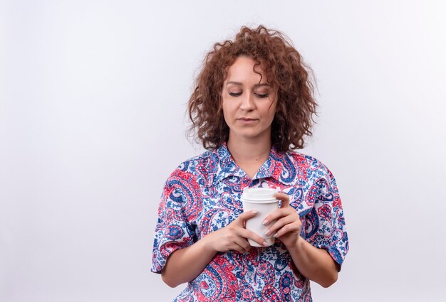 Junge Frau mit dem kurzen lockigen Haar im bunten Hemd, das Kaffeetasse hält, die es mit dem traurigen Ausdruck betrachtet, der über weißer Wand steht