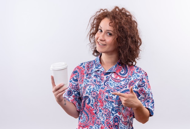 Junge Frau mit dem kurzen lockigen Haar im bunten Hemd, das Kaffeetasse hält, die mit dem Finger zeigt, der über weißer Wand lächelnd steht