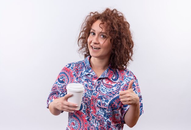 Junge Frau mit dem kurzen lockigen Haar im bunten Hemd, das Kaffeetasse hält lächelnd zeigt Daumen hoch stehend über weißer Wand