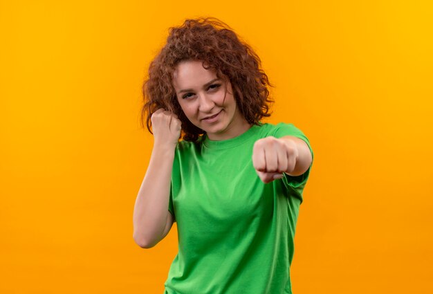 Junge Frau mit dem kurzen lockigen Haar im grünen T-Shirt, das Faust ballt, die wie Boxer aufstellt, der über orange Wand steht