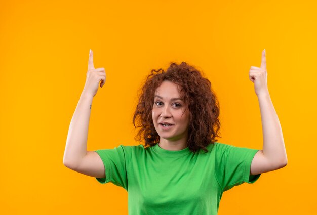 Junge Frau mit dem kurzen lockigen Haar im grünen T-Shirt, das lächelnd zeigt und mit den Fingern oben zeigt, die große Idee stehen