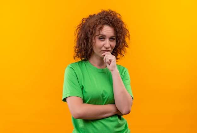 Junge Frau mit dem kurzen lockigen Haar im grünen T-Shirt, das mit nachdenklichem Ausdruck auf Gesicht denkend steht über orange Wand schaut