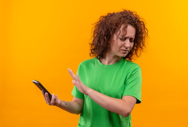 Junge Frau mit dem kurzen lockigen Haar im grünen T-Shirt, das Smartphone hält, das Verteidigungsgeste mit der anderen Hand gegen ihr Handy macht, das unzufrieden steht stehend