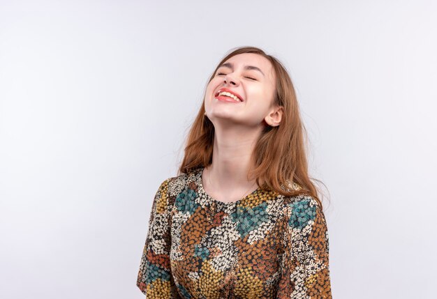 Junge Frau mit dem langen Haar, das buntes Kleid trägt, das mit geschlossenen Augen steht, die über weiße Wand lachen