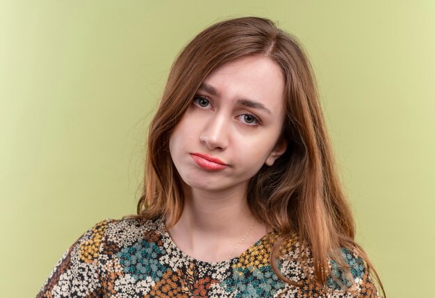 Junge Frau mit dem langen Haar, das buntes Kleid trägt, das sehr mit traurigem Ausdruck auf Gesicht steht, das über grüner Wand steht