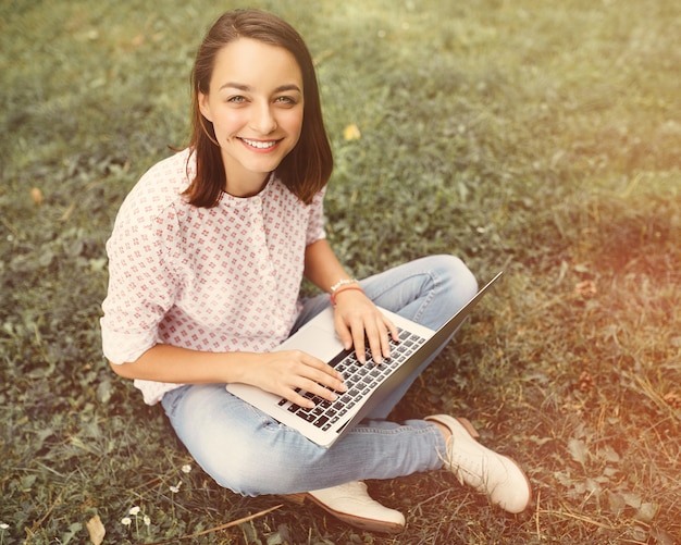 Junge Frau mit dem Laptop, der auf grünem Gras sitzt