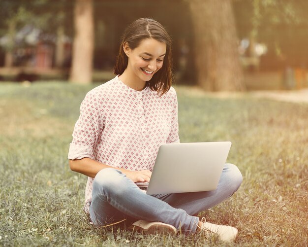 Junge Frau mit dem Laptop, der auf grünem Gras sitzt