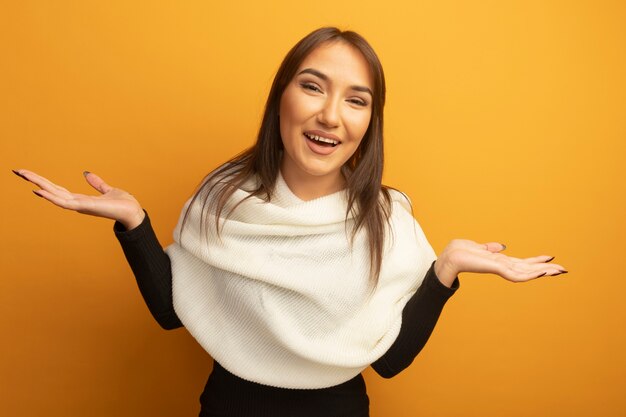 Junge Frau mit dem weißen Schal, der vorne lächelnd spreizende Arme zur Seite steht, die über orange Wand stehen