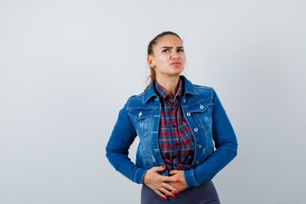 Junge Frau mit den Händen auf dem Bauch in kariertem Hemd, Jeansjacke und auf der Suche nach Schmerzen, Vorderansicht.