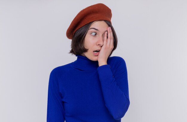 Junge Frau mit den kurzen Haaren im blauen Rollkragenpullover, der Baskenmütze trägt, die beiseite geschockt schaut, bedeckt ein Auge mit Hand, die über weißer Wand steht