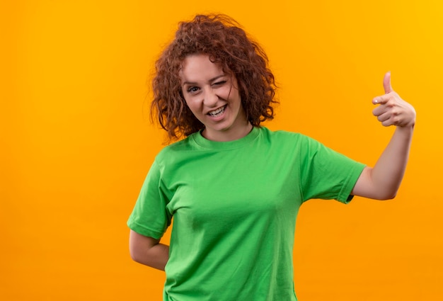 Kostenloses Foto junge frau mit den kurzen lockigen haaren im grünen t-shirt glücklich und positiv zwinkert, die daumen hoch stehend über orange wand zeigt
