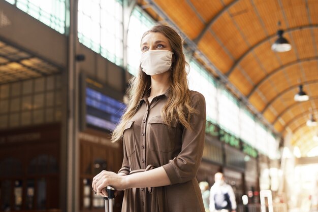 Junge Frau mit einem Koffer, der eine Gesichtsmaske trägt und an einem Bahnhof wartet - COVID-19