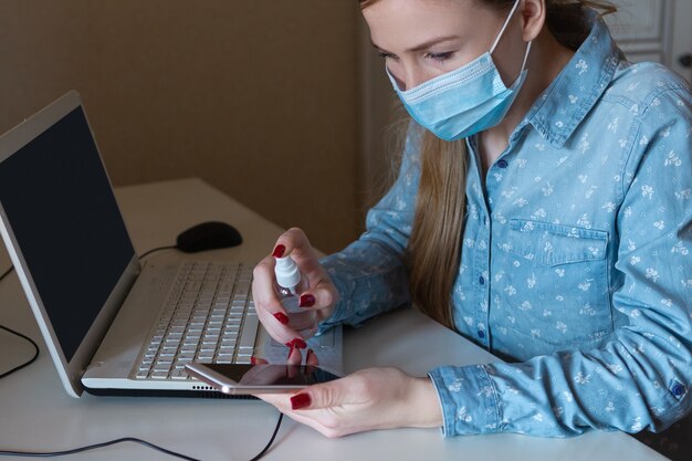 Junge frau mit gesichtsmaske desinfiziert geräteoberflächen an ihrem arbeitsplatz