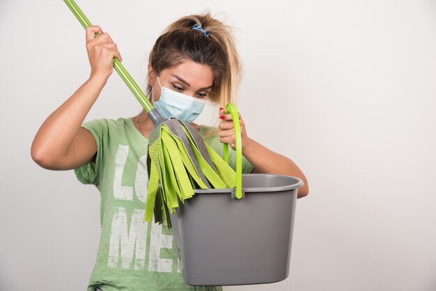 Junge Frau mit Gesichtsmaske, die Mopp auf weißer Wand betrachtet.