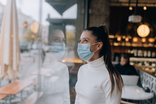 Junge Frau mit Gesichtsmaske steht vor Fenstern im Café