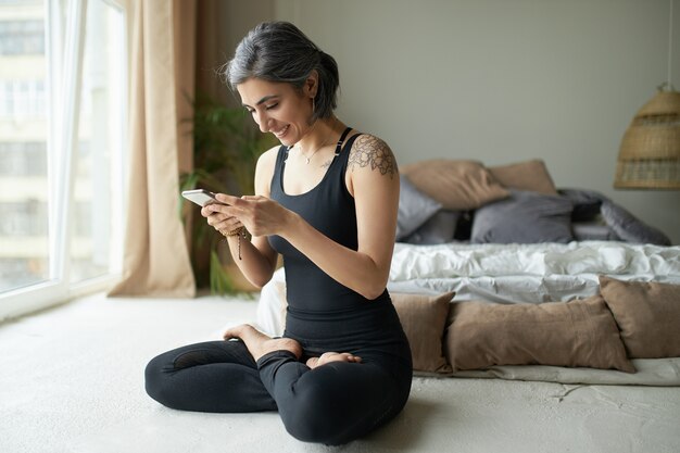 Junge Frau mit grauem Haar und Tätowierung, die Yoga praktiziert und das Smartphone benutzt