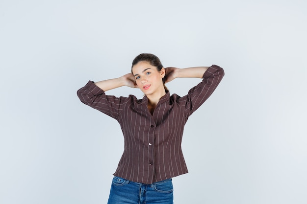 Junge Frau mit Händen hinter dem Kopf in gestreiftem Hemd, Jeans und selbstbewusstem Blick. Vorderansicht.