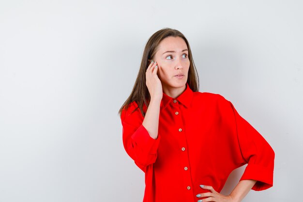 Junge Frau mit Hand in der Nähe von Gesicht und Taille in roter Bluse und nachdenklich aussehend, Vorderansicht.