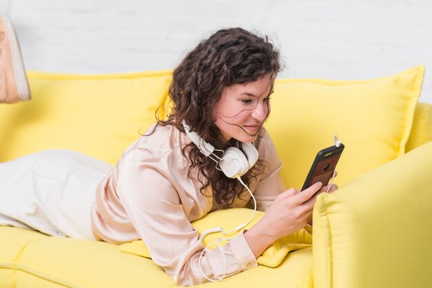 Junge Frau mit Kopfhörer um ihren Hals, der auf gelbem Sofa unter Verwendung des Smartphone liegt