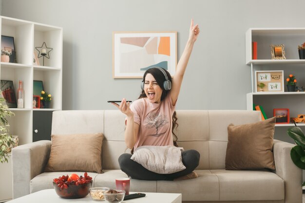 Junge Frau mit Kopfhörern, die Telefonanschlüsse auf dem Sofa hinter dem Couchtisch im Wohnzimmer hält