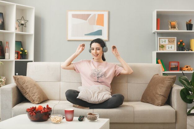 Junge Frau mit Kopfhörern mit Keksen beim Yoga auf dem Sofa hinter dem Couchtisch im Wohnzimmer