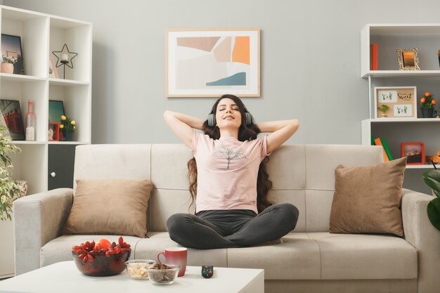 Junge Frau mit Kopfhörern sitzt auf dem Sofa hinter dem Couchtisch im Wohnzimmer