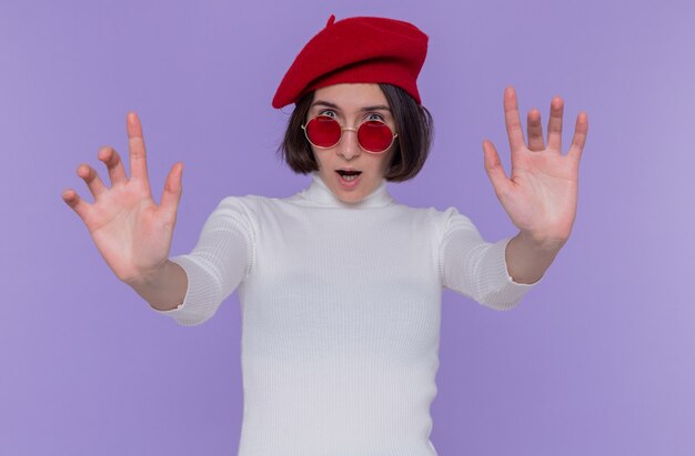 Junge Frau mit kurzen Haaren im weißen Rollkragenpullover, der Baskenmütze und rote Sonnenbrille trägt, die besorgte und verwirrte Front betrachten, die Stoppgeste mit den Händen macht, die über blauer Wand stehen