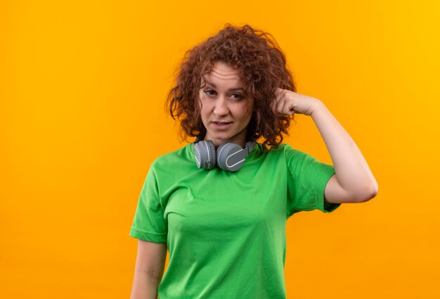 Junge Frau mit kurzen lockigen Haaren im grünen T-Shirt mit Kopfhörern, die verwirrt schauen und mit dem Finger ihre Schläfe zeigen über orange Wand stehen