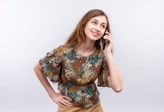 Junge Frau mit langen Haaren, die buntes Kleid lächelnd beim Sprechen auf Handy tragen, das über weißer Wand steht