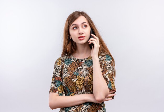 Junge Frau mit langen Haaren, die buntes Kleid lächelnd beim Sprechen auf Handy tragen, das über weißer Wand steht