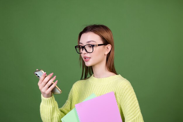 Junge Frau mit perfektem natürlichem Make-up braune große Lippen in lässigem Pullover auf grüner Wandstudentin in Gläsern und Notebooks mit Handy