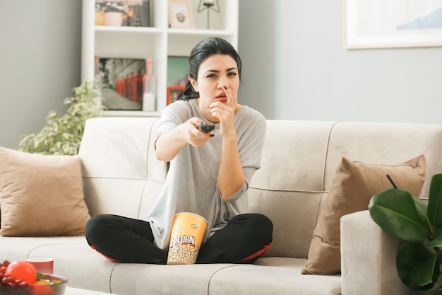 Junge Frau mit Popcorn-Eimer hält die TV-Fernbedienung zur Kamera auf dem Sofa hinter dem Couchtisch im Wohnzimmer