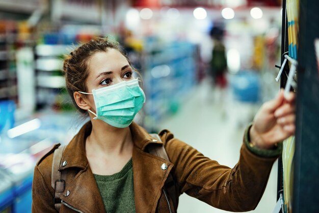 Junge Frau mit Schutzmaske im Gesicht, die während der Coronavirus-Pandemie im Supermarkt kauft