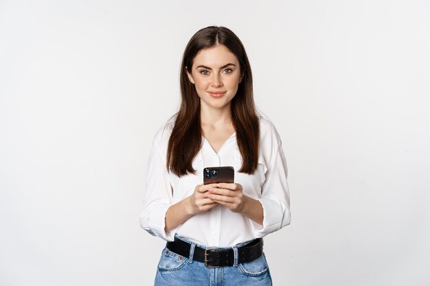 Junge Frau mit Smartphone, lächelnd und in die Kamera schauend, mit Handy-App, Mobilfunktechnologie und Online-Shopping-Konzept, weißer Hintergrund