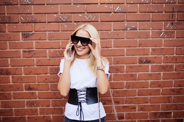 Junge Frau mit Sonnenbrille lachend, während sie am Handy neben brauner Backsteinmauer im Freien am sonnigen Tag spricht