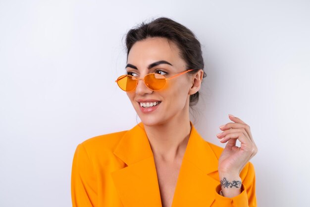 Junge Frau mit trendiger, stylischer Brille und einer leuchtend orangefarbenen, übergroßen Jacke auf weißem Hintergrund