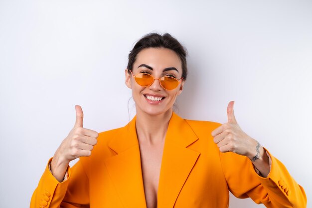 Junge Frau mit trendiger, stylischer Brille und einer leuchtend orangefarbenen, übergroßen Jacke auf weißem Hintergrund