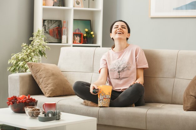 Junge Frau mit TV-Fernbedienung sitzt auf dem Sofa hinter dem Couchtisch im Wohnzimmer