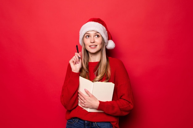 Junge Frau mit Weihnachtsmütze hält einen Notizblock in den Händen und beißt einen Stift, der die Liste der Geschenke oder Kinder mit gutem Benehmen einzeln auf einer roten Wand überprüft.,
