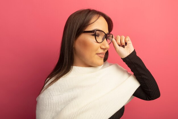 Junge Frau mit weißem Schal in den Gläsern, die beiseite schauen und ihre Brille berühren, die über rosa Wand steht