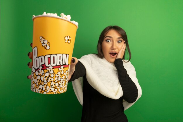 Junge Frau mit weißem Schal zeigt Eimer mit Popcorn erstaunt und überrascht