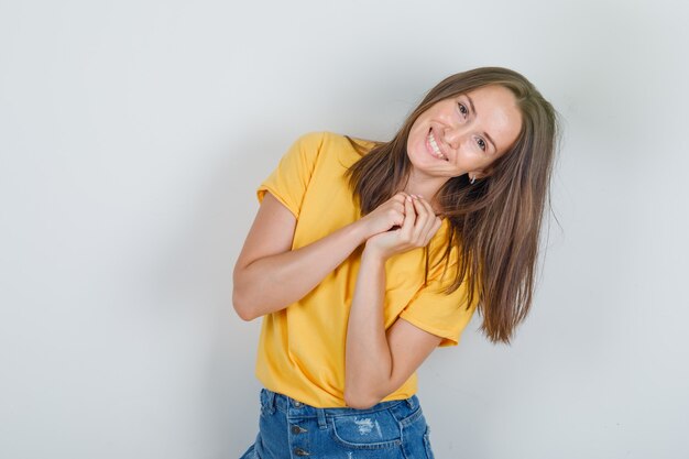 Junge Frau posiert wie um einen Gefallen in gelbem T-Shirt, Shorts bitten und fröhlich aussehen